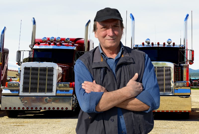 A male truck driver wearing a black vest over a blue shirt and a black baseball cap stands in front of two semi trucks.