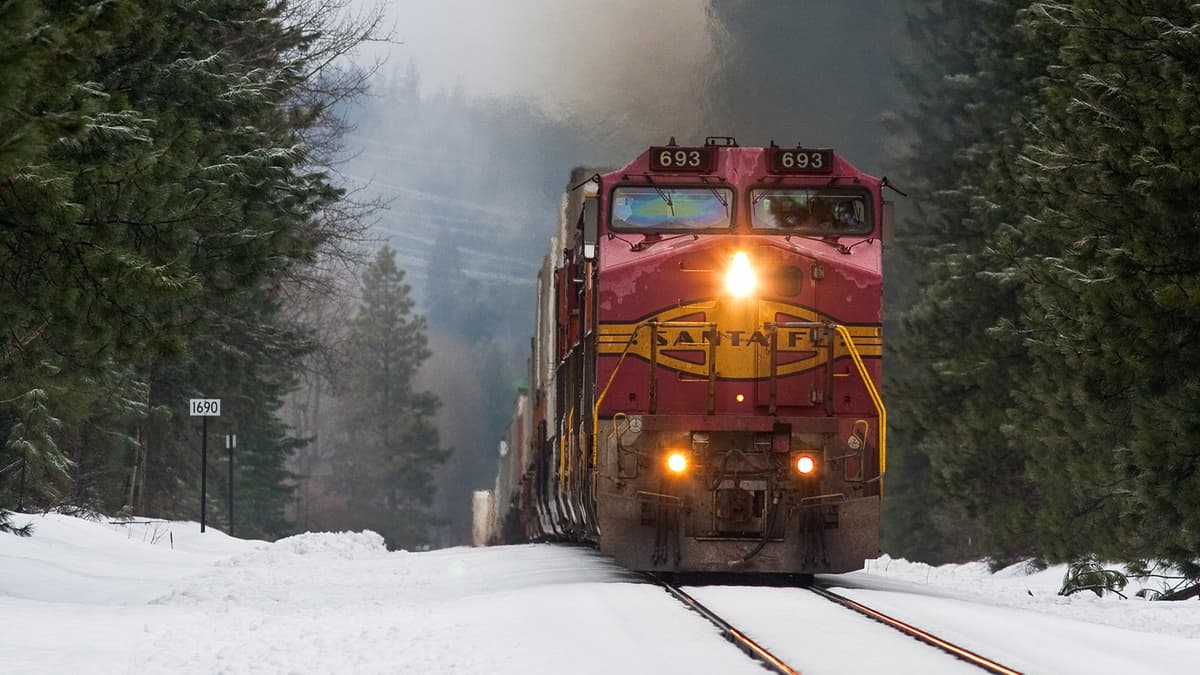 A photograph of a train with a headlight on. There is snow on the ground.