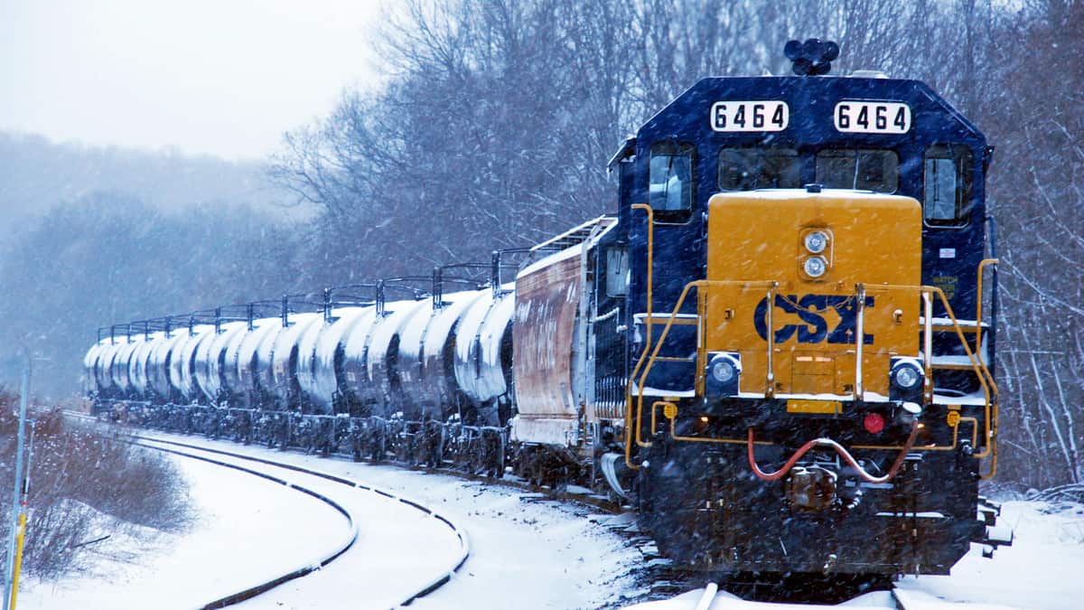 A photograph of a train hauling tank cars in winter. There is snow on the ground.
