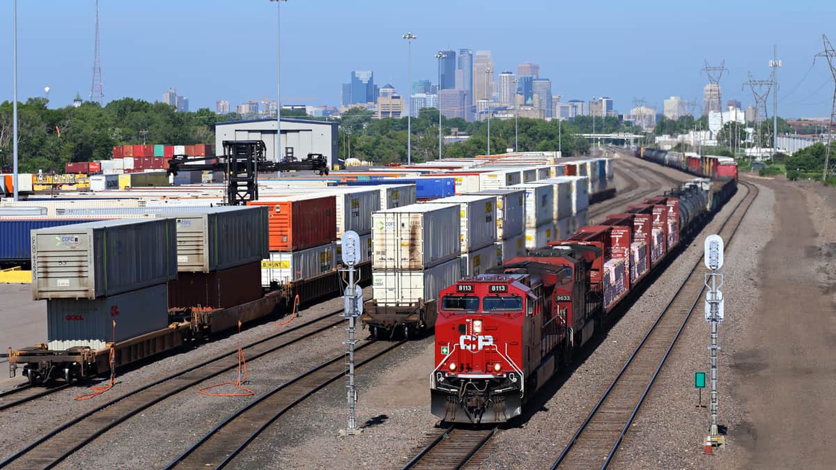 A photograph of a train in a rail yard. Intermodal containers are at the rail yard, sitting nearby.