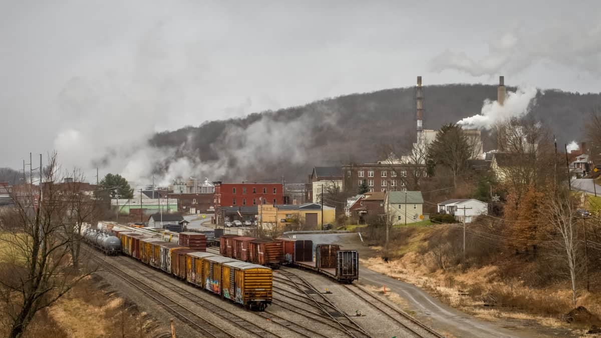 A photograph of boxcars and tank cars parked on rail tracks. The tracks are next to some town buildings. A tree-covered mountain is behind the buildings.