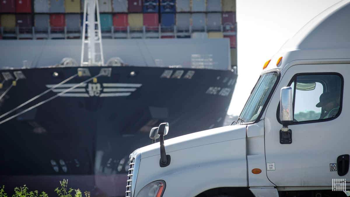 Driver in truck at dock with ship in background.