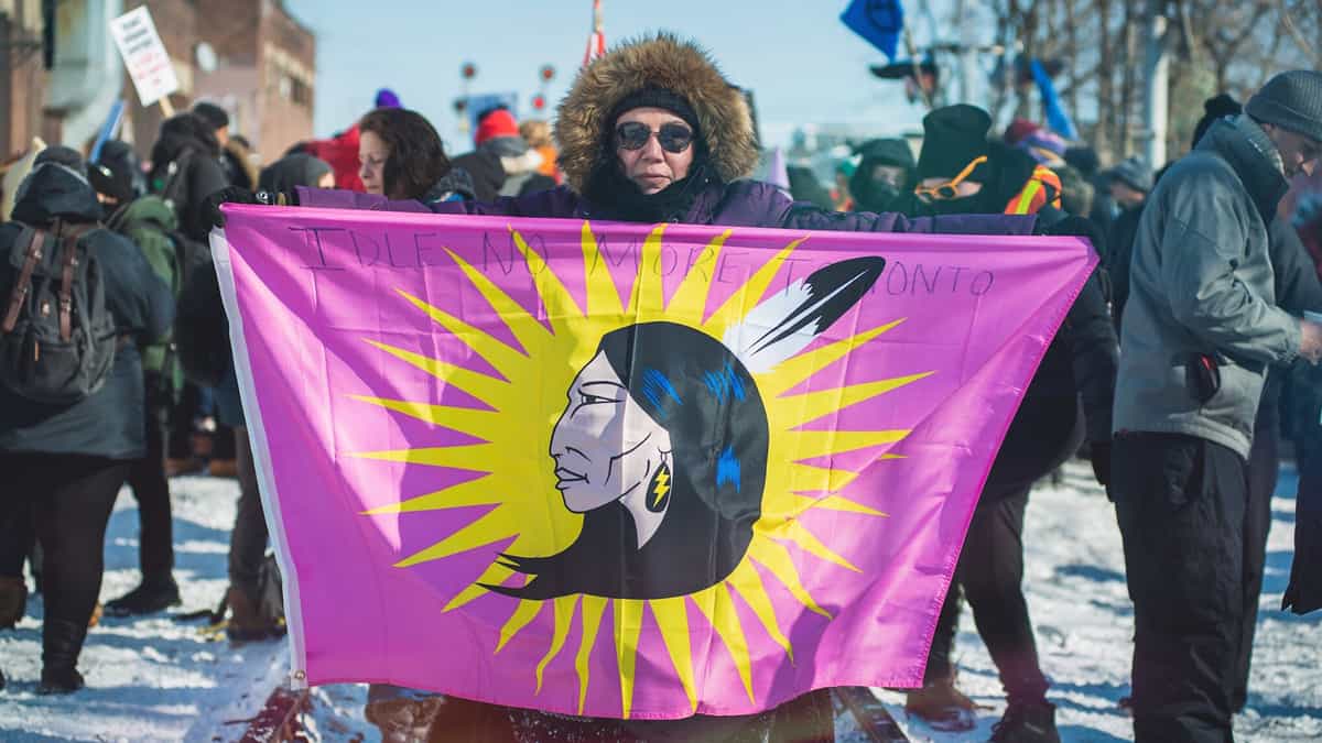 A photograph of a person holding a flag with the image of a First Nation person on it. There is a crowd of people behind the person.