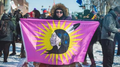 A photograph of a person holding a flag with the image of a First Nation person on it. There is a crowd of people behind the person.