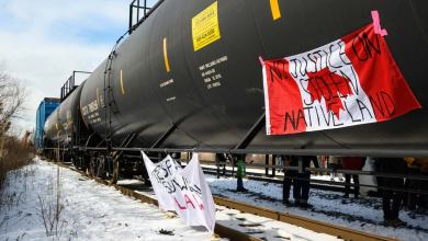 A photograph of tank cars on a rail track. One of the tank cars has a Canadian flag hanging from it. Written on the flag are the words, "no justice on stolen native land."