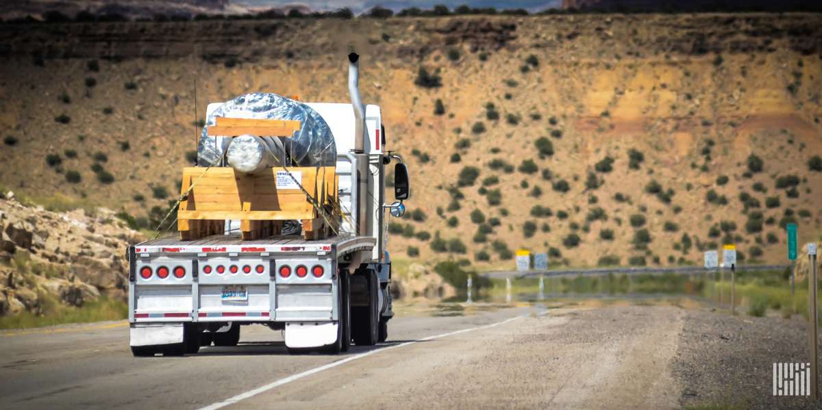 A white commercial truck with a flatbed trailer with wooden cargo strapped to it, seen from the rear as it travels on a highway.