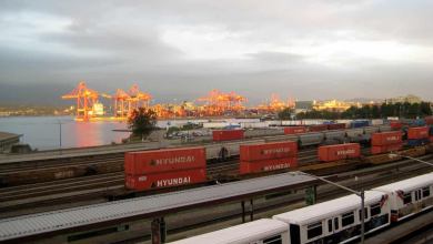 A photograph of a rail yard. There are intermodal containers and rail cars There are loading cranes at a port in the background.