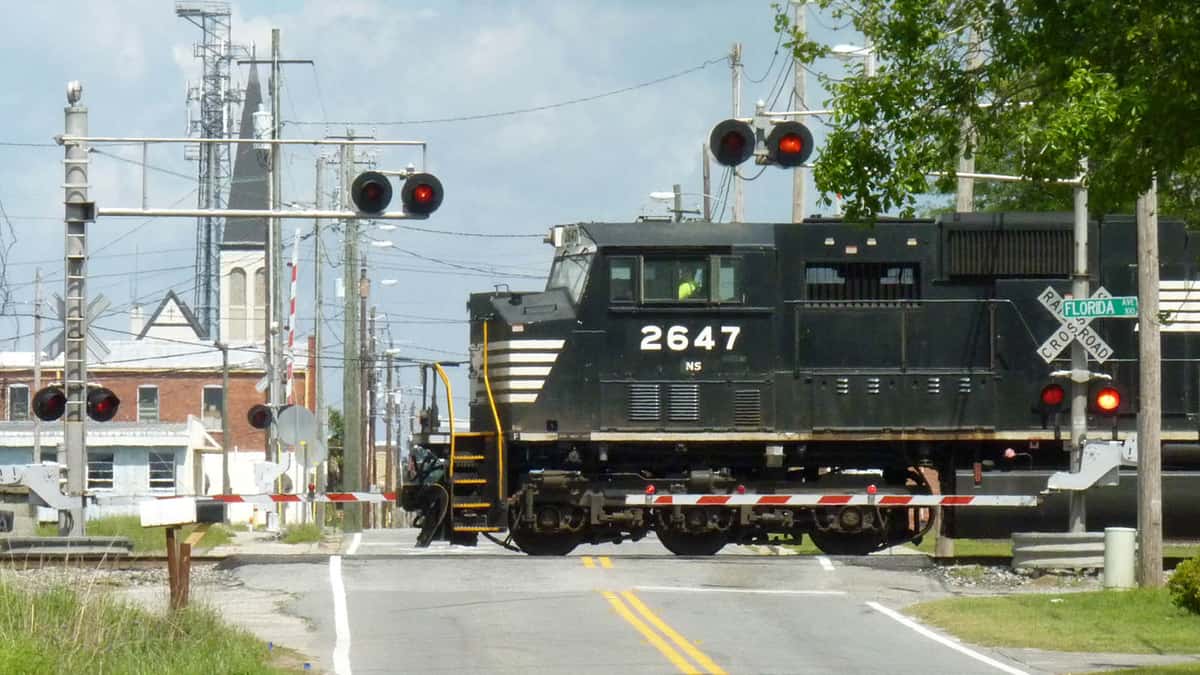 A photograph of a train on railroad tracks crossing a town road. There are signs and signals around the train for drivers and pedestrians.