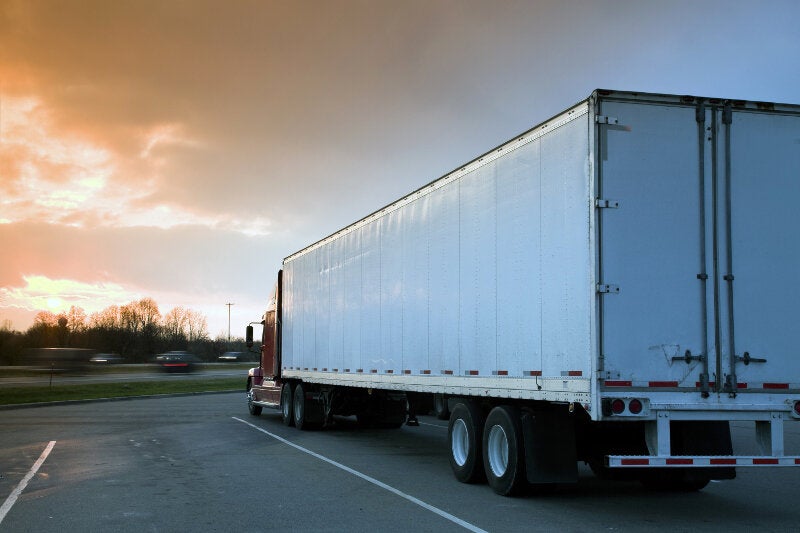 A red and white tractor trailer is parked in a parking lot with a sunset behind it.