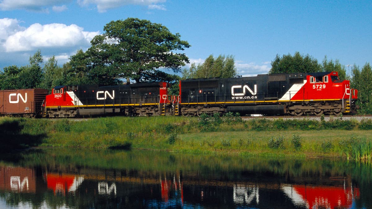 A photograph of a train. It is running next to a pond and a reflection of the train is on the pond's surface.