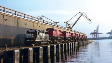A photograph of a train on the dock of a port. The train is next to the water.