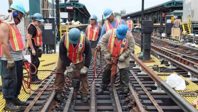 A photograph of workers performing work on a rail track near a passenger railway station.