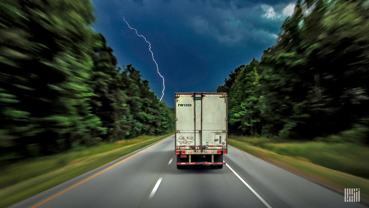 Tractor-trailer heading down highway with lightning in the sky ahead.