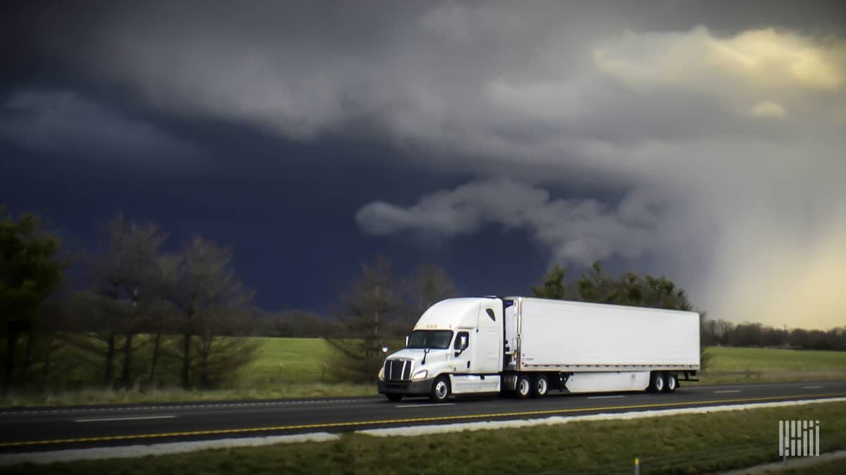 Tractor-trailer heading down highway with thunderstorm cloud in background.