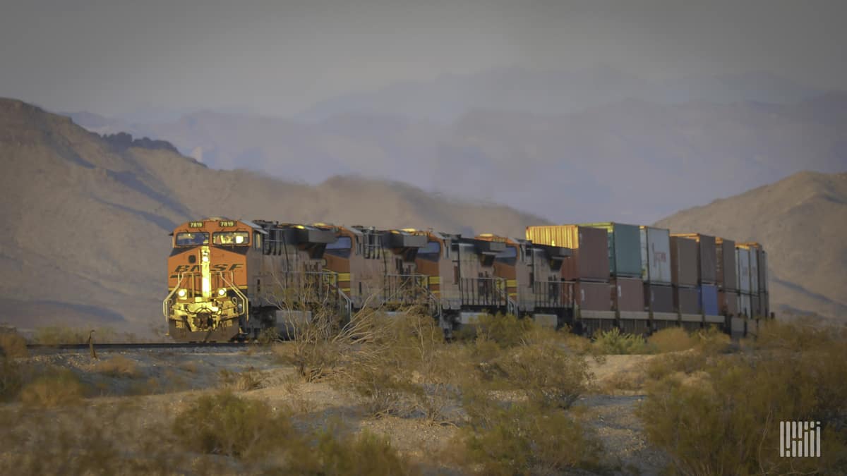 A train hauling railcars through a field. There is a mountain range in the background.