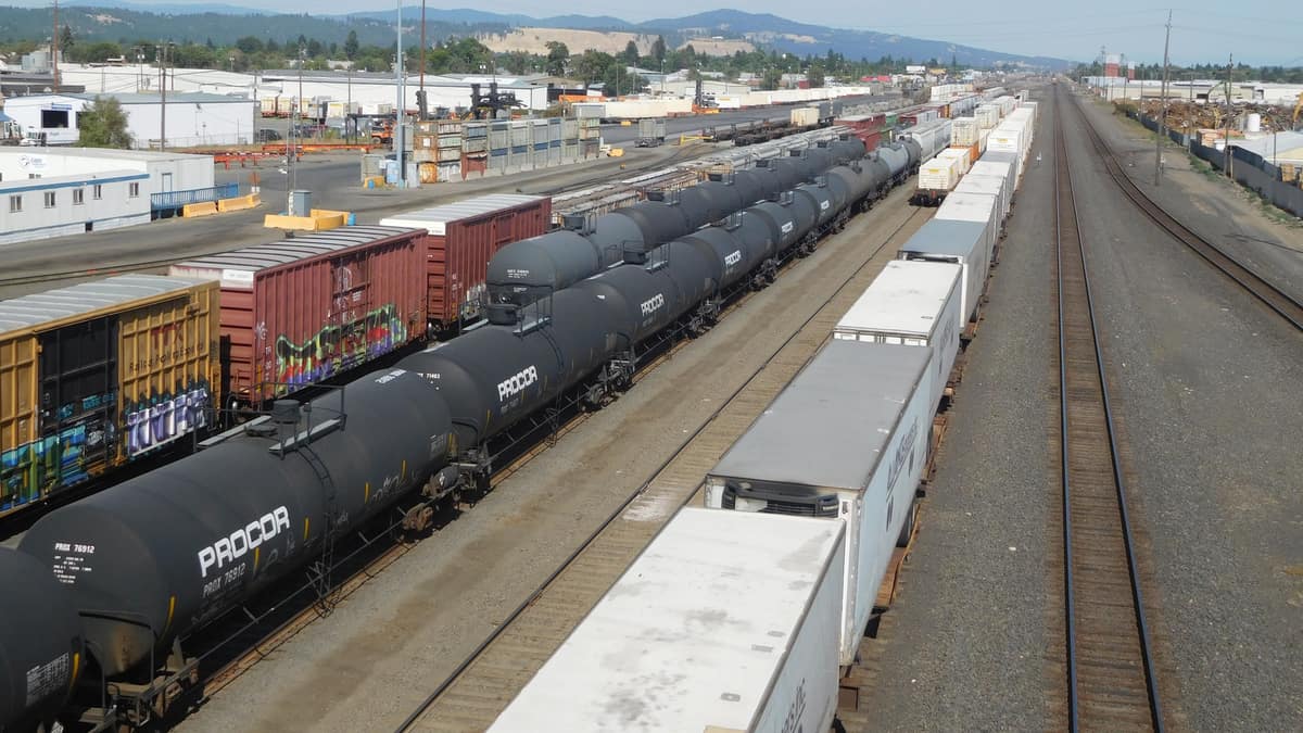 A photograph of a line of boxcars, a line of tank cars and a line of intermodal containers, all parked at a rail yard.