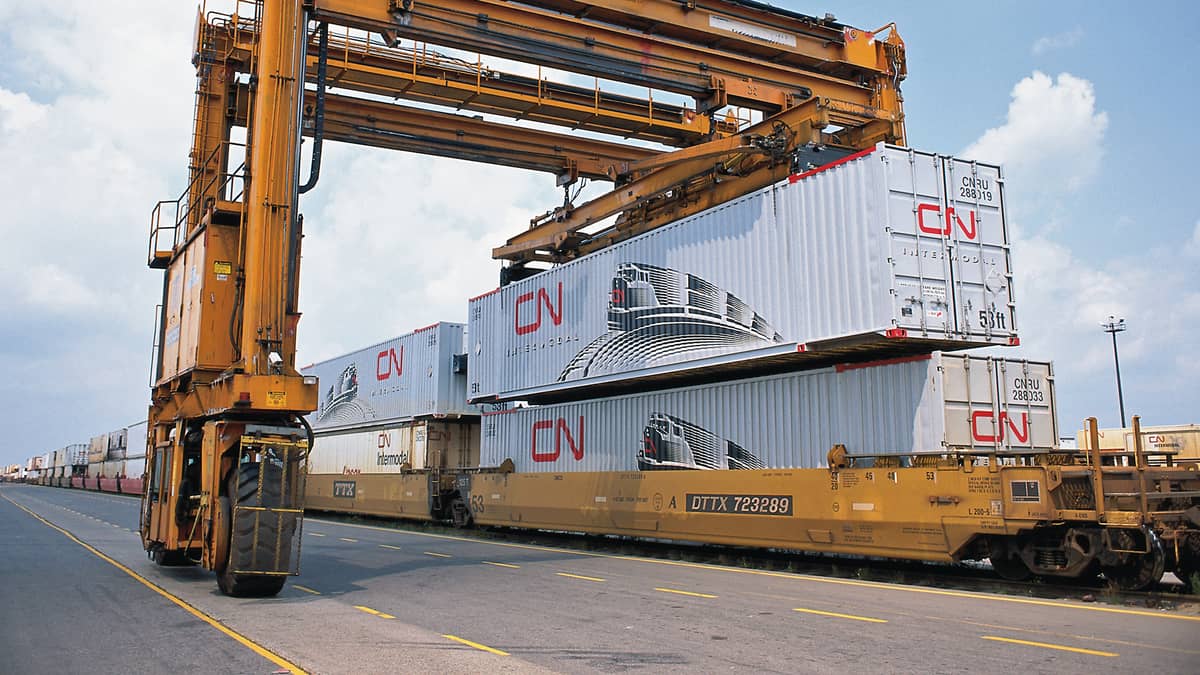 A photograph of tall cranes lifting an intermodal container. That container is stacked on top of another container.