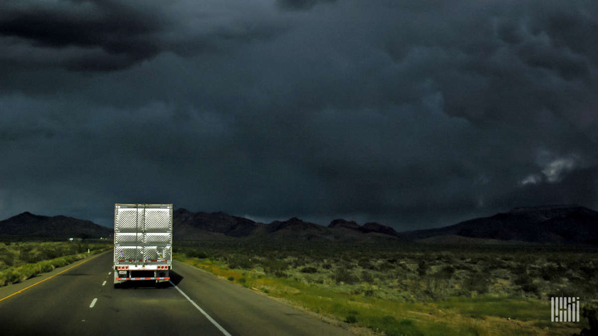 Tractor-trailer heading down highway with dark thunderstorm ahead.