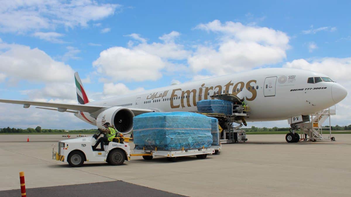 A white Emirates plane on tarmac unloading pallets of freight, pulled by a tug under bright blue sky. Emirates is running passenger freighters to Rickenbacker airport in Ohio.