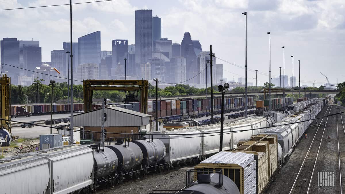 A photograph of trains at a rail yard. There are tall city buildings in the background.