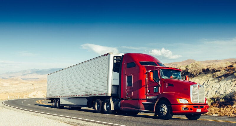 A red semi-truck pulling a white trailer with a black temperature control unit drives around the a curve on a desert highway.