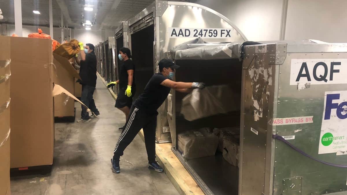 Warehouse workers unload metal containers filled with small mail envelopes and parcels that arrived by air. Cargo Force does sort and prep work for the USPS.