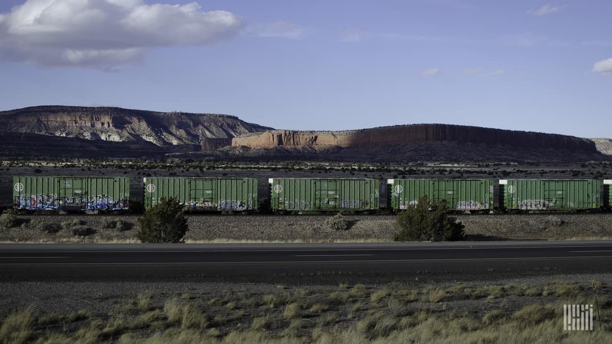 A photograph of a train of boxcars traveling through a desert-like field. There is a mountain range in the background.