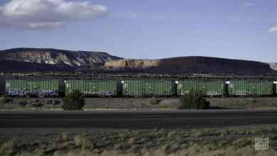 A photograph of a train of boxcars traveling through a desert-like field. There is a mountain range in the background.