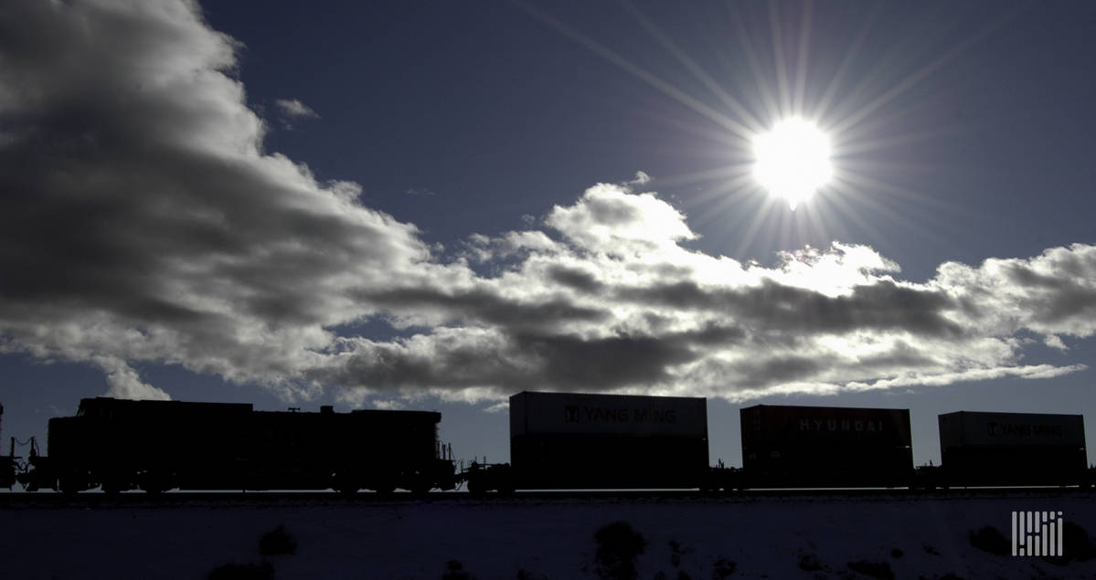 A photograph of a train carrying intermodal containers across a field. The sun is bright in the sky above the train.