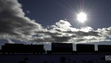 A photograph of a train carrying intermodal containers across a field. The sun is bright in the sky above the train.