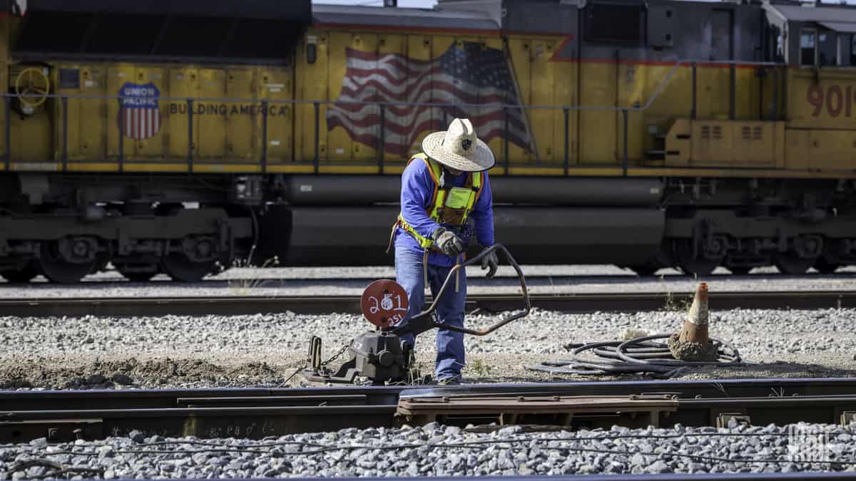 A photograph of a worker fixing track in a rail yard. There is a parked train behind him.