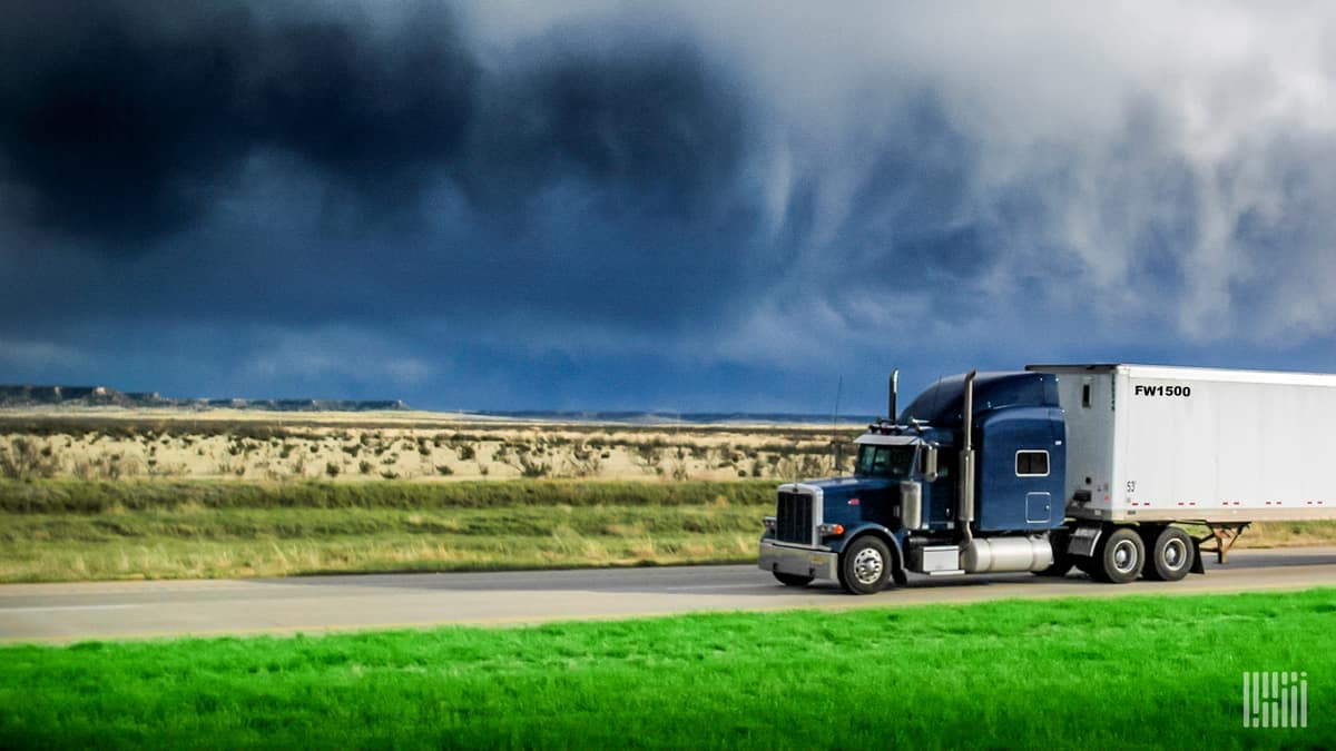 Tractor-trailer heading down highway with dark storm cloud across the sky.