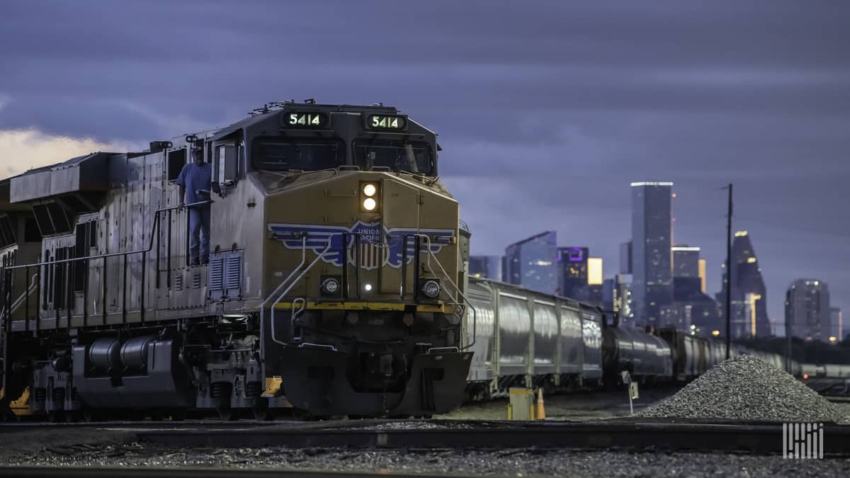 A photograph of a freight train travelling in front of tall city buildings.