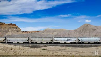 A photograph of a train with hoppers. The train is crossing a desert-like landscape.