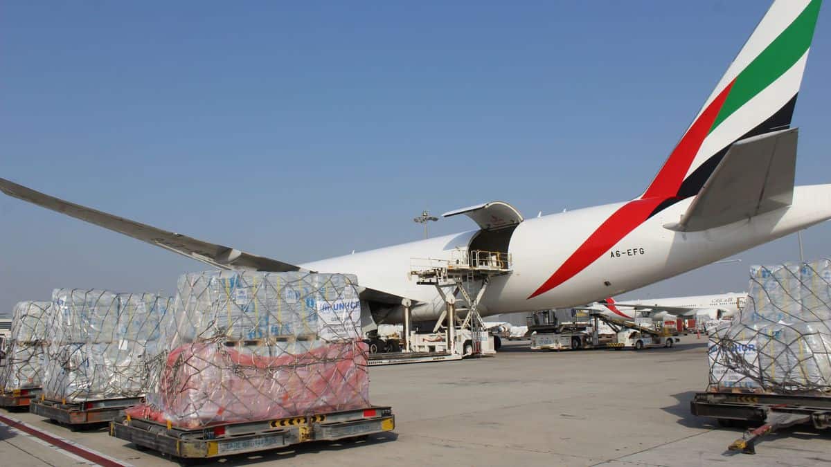 Pallets of humanitarian cargo sit outside next to a big plane waiting to be loaded on a sunny day.