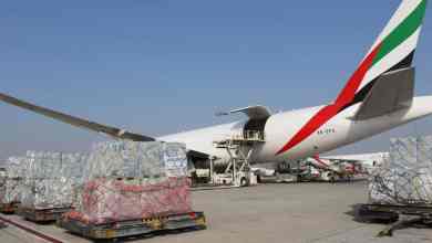 Pallets of humanitarian cargo sit outside next to a big plane waiting to be loaded on a sunny day.