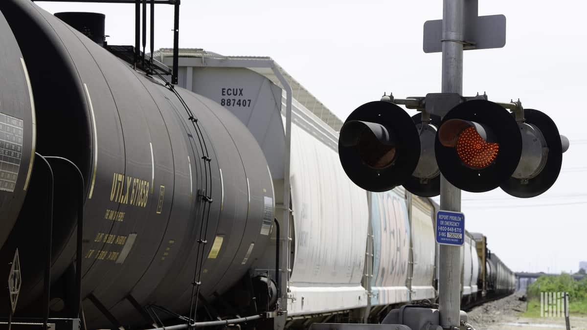 A photograph of a tank car and a hopper traveling by a railroad crossing.