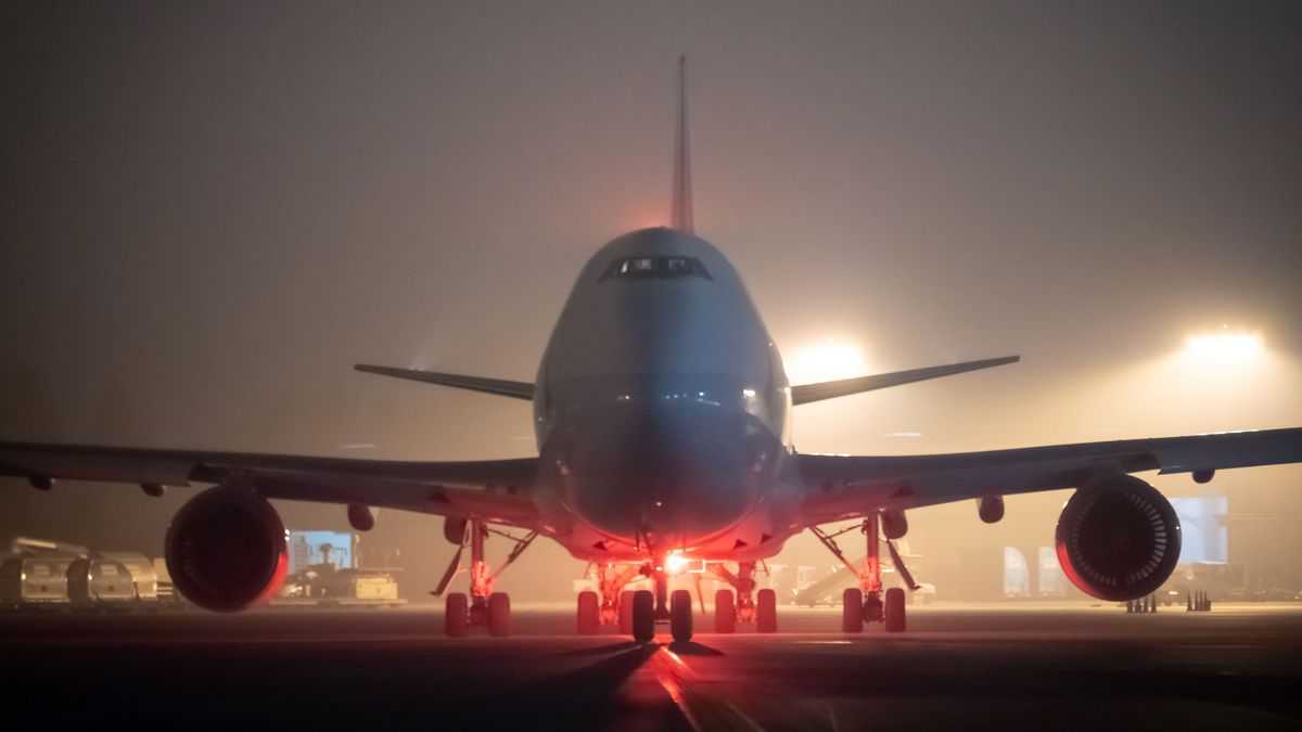 A cargo jumbo jet facing the camera silhouetted in night sky with smoke from fires creating red background.