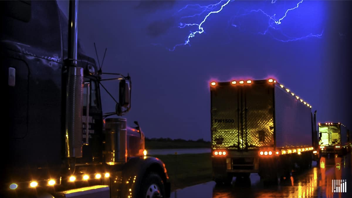 Tractor-trailers heading down highway in the rain, with lightning across the sky.