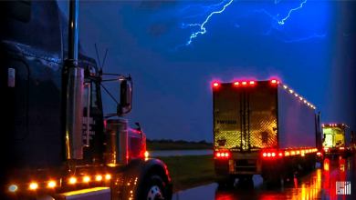 Tractor-trailers heading down highway in the rain, with lightning across the sky.