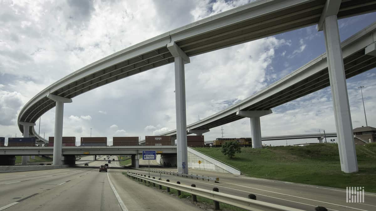 A photograph of a train hauling intermodal containers by a highway overpass.