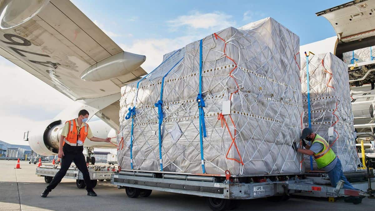 Big pallet of shrink-wrapped cargo being rolled under the wing of a cargo plane on a sunny day.