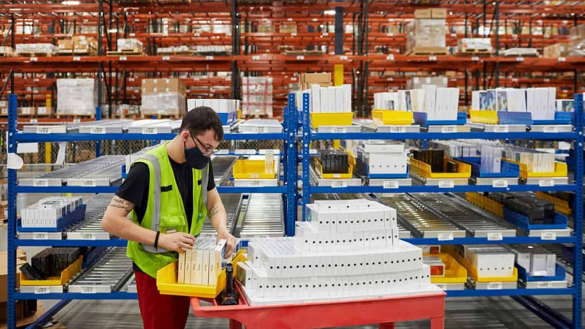 A man wearing yellow vest moves a cart loaded with small boxes inside a big Apple warehouse.