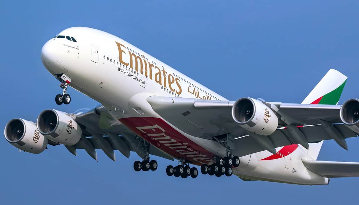 A giant white Emirates plane with four engines and wheels down comes in for a landing behind a blue sky.