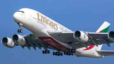 A giant white Emirates plane with four engines and wheels down comes in for a landing behind a blue sky.