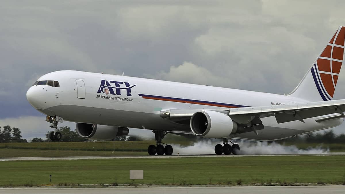 A large white plane with red tail touches down on runway. ATI is lettering.