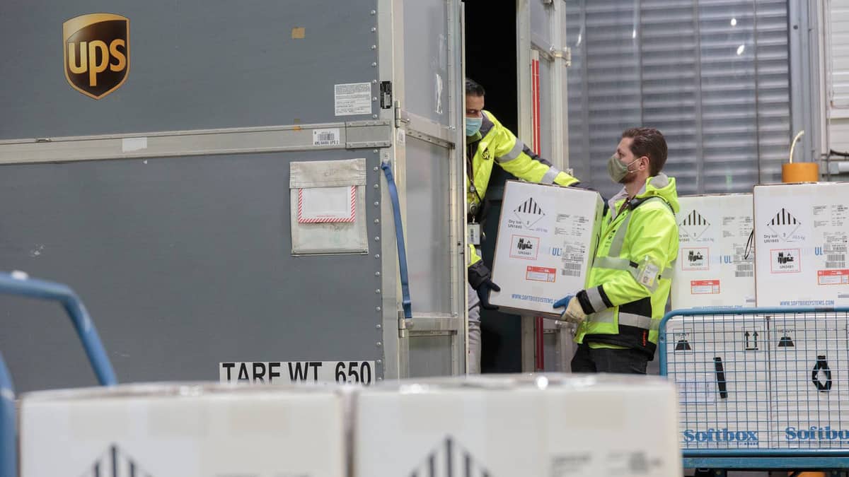 UPS drivers wearing yellow raincoats unload boxes of vaccine from a van.