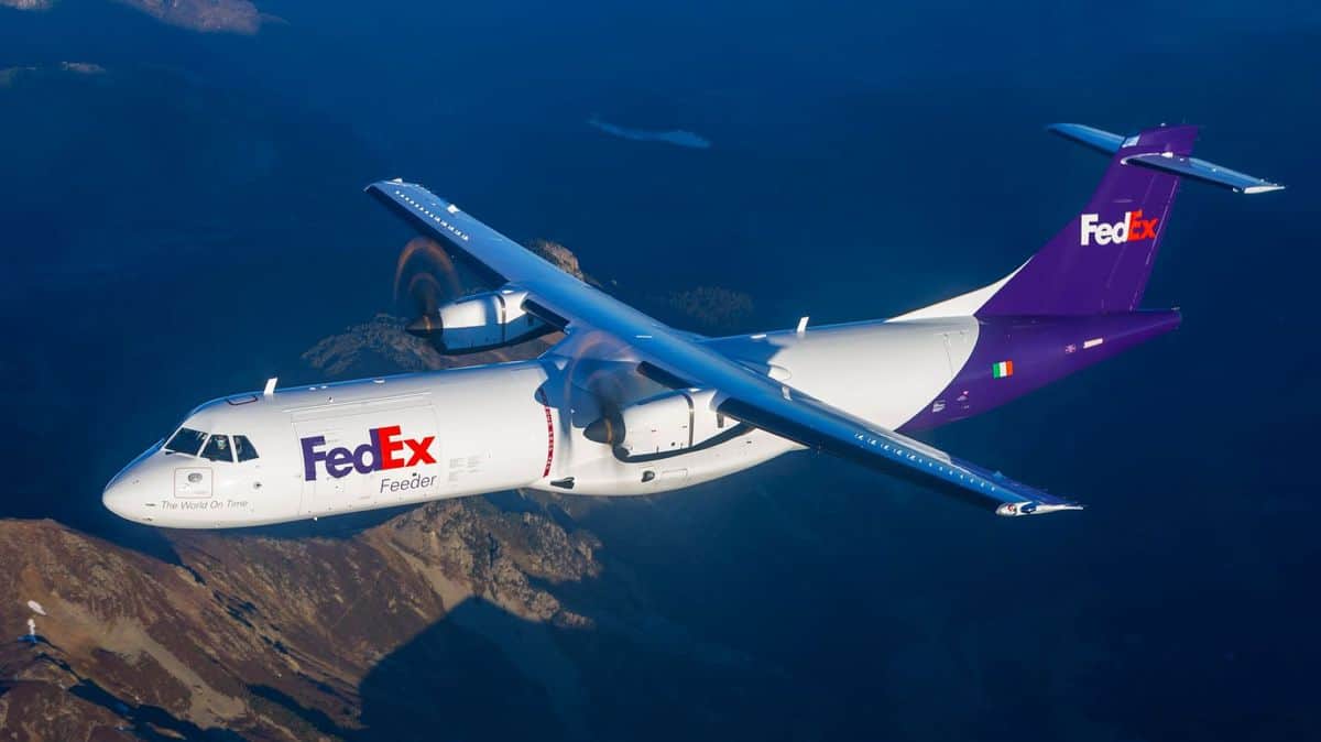 A white FedEx propeller plane flies on clear day, with ground visible below.
