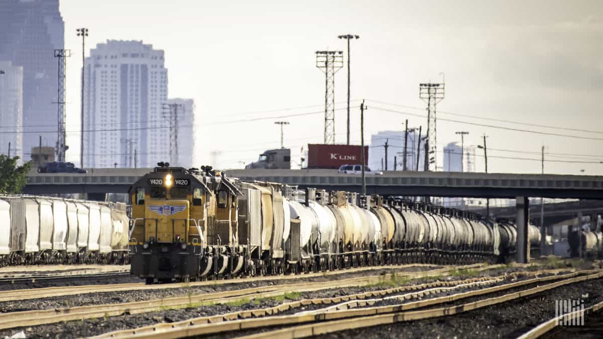 A photograph of a two trains sitting in a rail yard. Tall buildings are behind the rail yard.