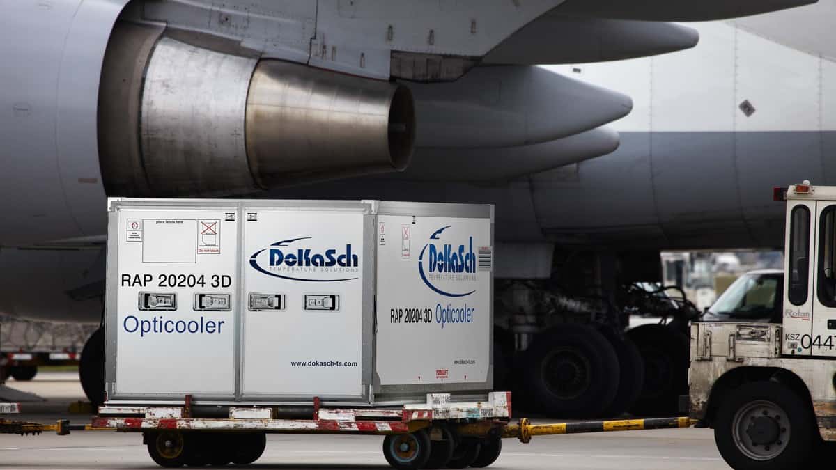 A large cooler on wheels for medicine transport sits on tarmac beneath an airplane wing. Air transport is commonly used to ship medicines.